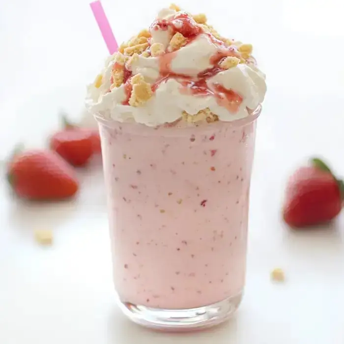 A creamy pink drink topped with whipped cream, cookie crumbles, and berry syrup, sitting with fresh strawberries and a straw.