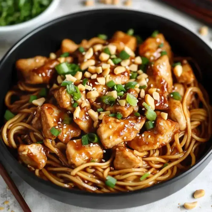 Close-up of a black bowl filled with noodles, chicken pieces, chopped green onions, peanuts, and sesame seeds in rich sauce.