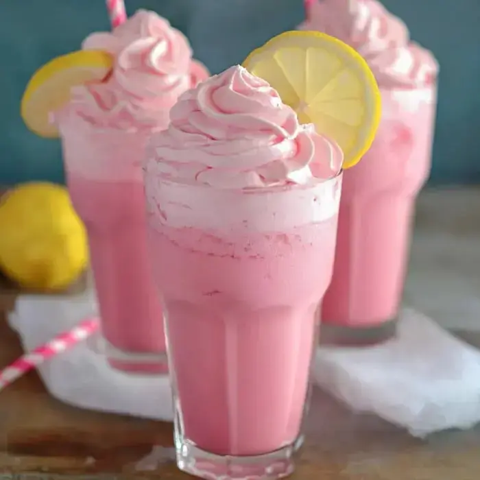 Three tall glasses filled with pink frothy drinks, topped with fluffy whipped cream, lemon slices and a colorful straw, sitting next to a lemon.