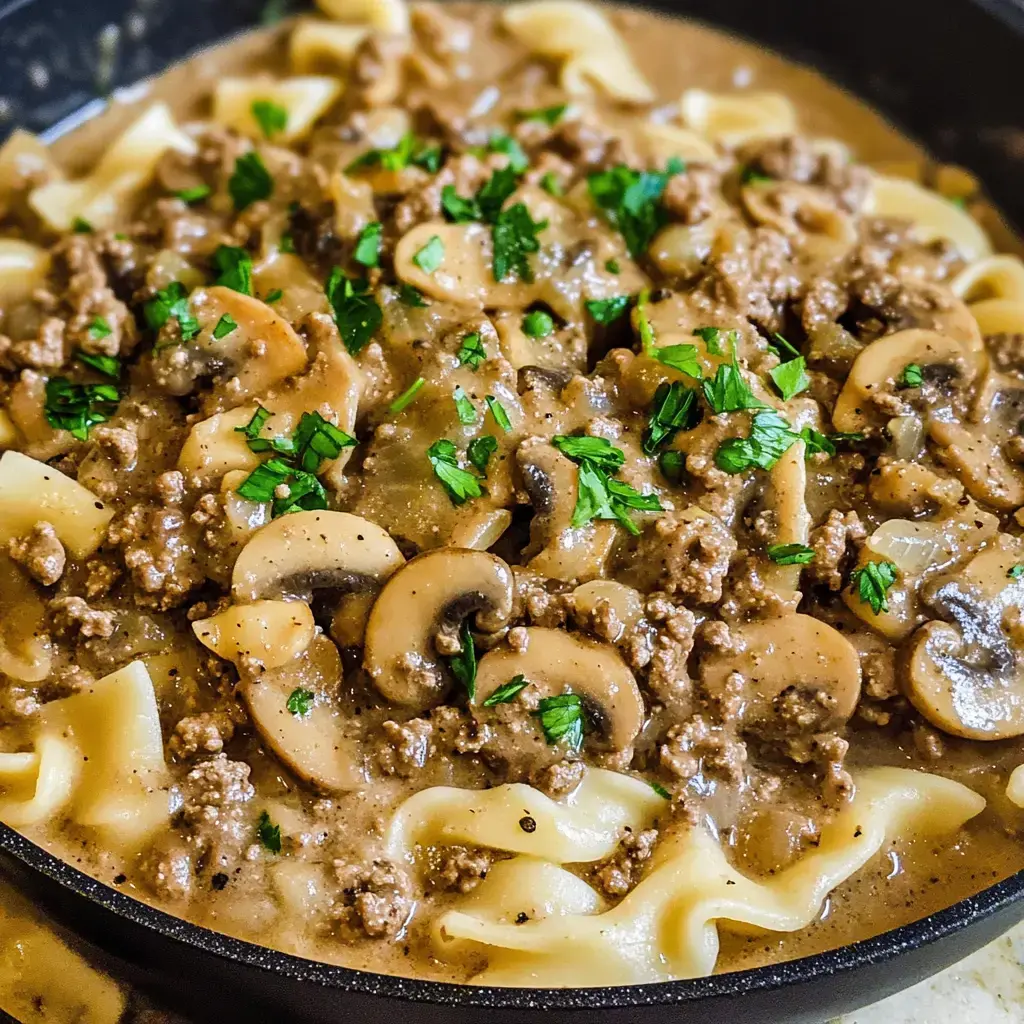 A frying pan containing egg noodles covered with a rich mixture of ground beef, sliced mushrooms, and scattered fresh parsley.
