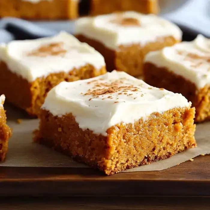 Pumpkin bars up close, with creamy frosting and a sprinkle of cinnamon, all on a wood background.