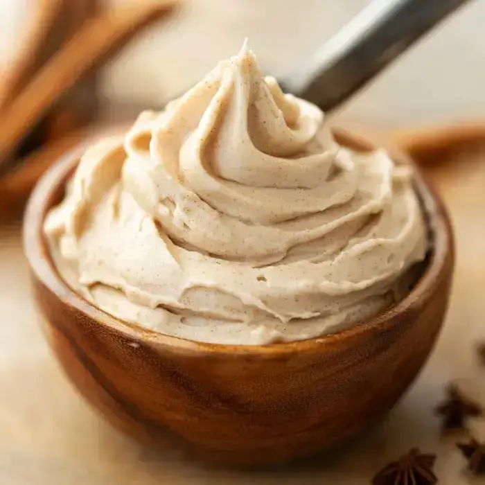 Silky light brown frosting twirled in a wooden bowl with cinnamon sticks scattered close by.
