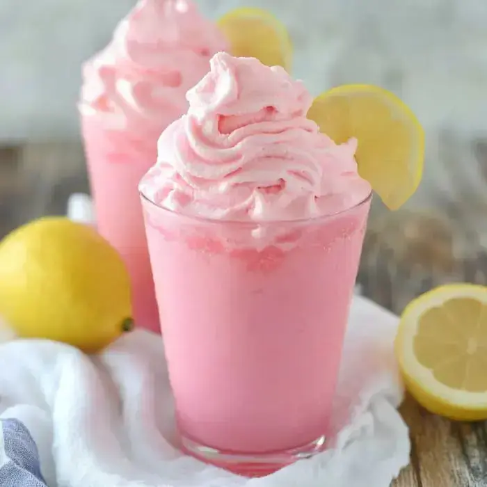 Two fun pink lemonade drinks with whipped tops and lemon wheels, surrounded by fresh lemons on a light cloth.