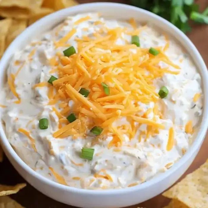 Bowlful of smooth dip, topped with cheddar shreds and green onions, hanging out with tortilla chips.