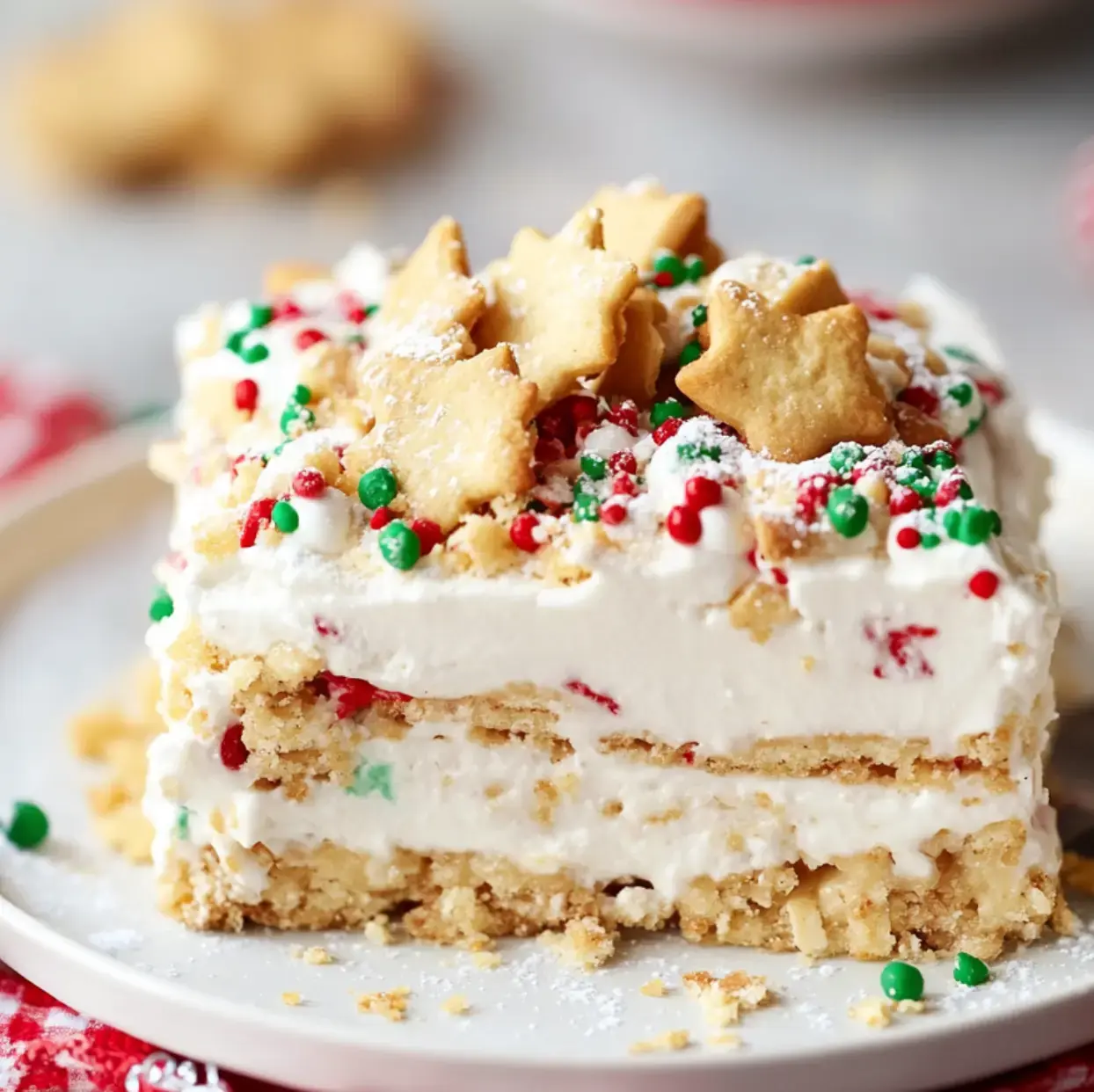 A piece of creamy layered treat with whipped topping, bright sprinkles, and shaped holiday cookies on a plate.