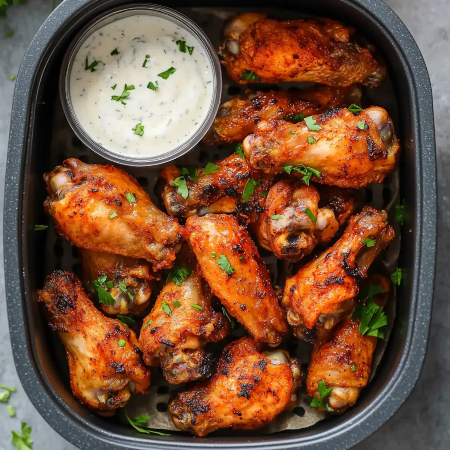 A plate of golden, seasoned chicken wings with fresh parsley sprinkled on top, next to a small container of dipping sauce.