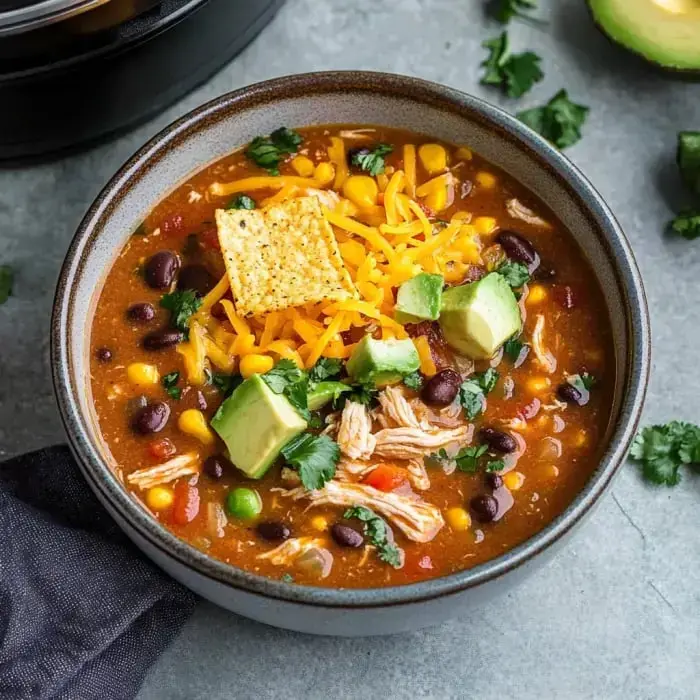 A comforting bowl of soup featuring juicy chicken, sweet corn, black beans, creamy avocado chunks, crispy tortilla chips, and shredded cheese with fresh cilantro.