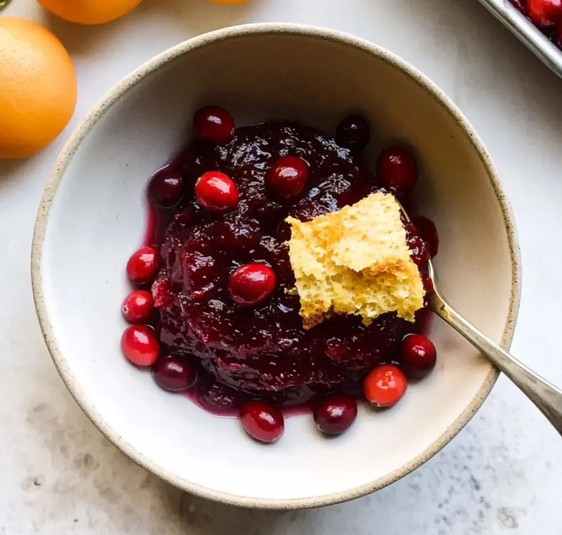 Bowl of cranberry sauce with fresh cranberries, a cornbread slice, and oranges in the background.
