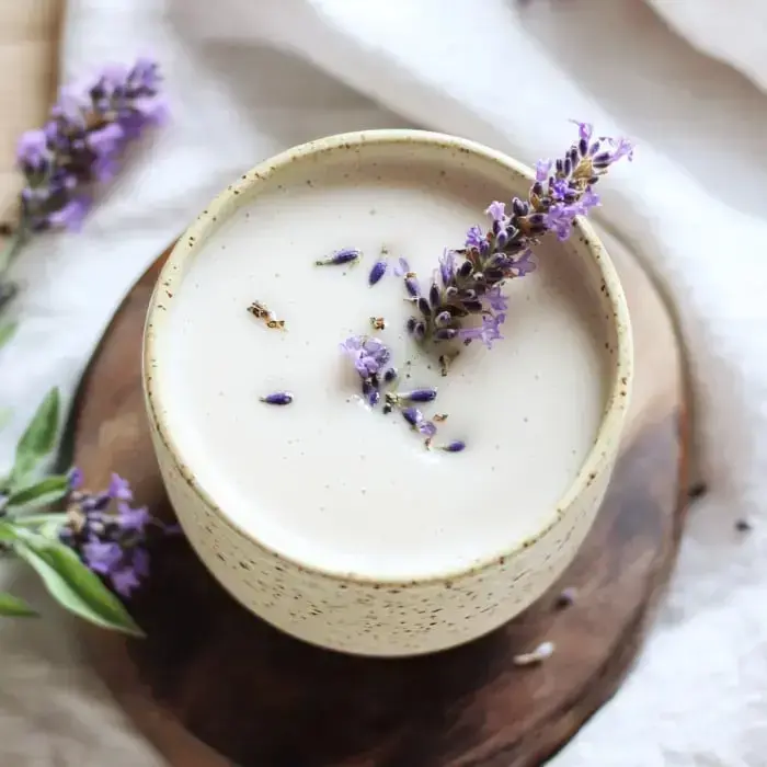 A creamy drink topped with fresh lavender sprigs in a speckled clay mug sitting on a wooden table surface.