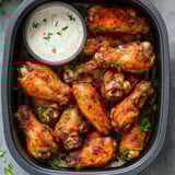 A plate of golden, seasoned chicken wings with fresh parsley sprinkled on top, next to a small container of dipping sauce.