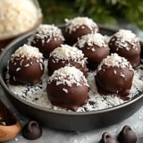 A plate of chocolate-coated coconut balls sprinkled with coconut flakes sits on a gray background.