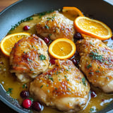 A view of golden chicken thighs topped with orange pieces and bright red berries in a flavorful sauce, presented in a cast iron pan.