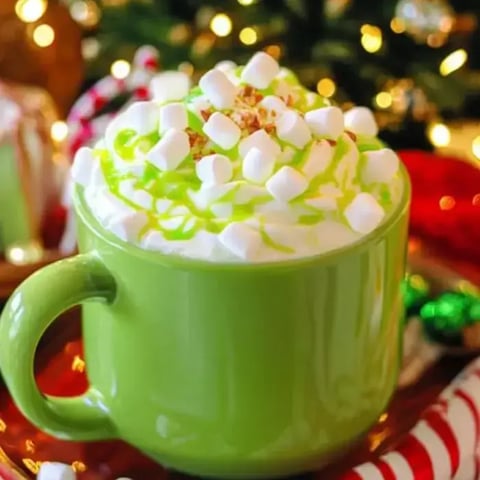 Bright green mug filled with whipped cream, marshmallows, and green drizzle, surrounded by festive holiday decor.
